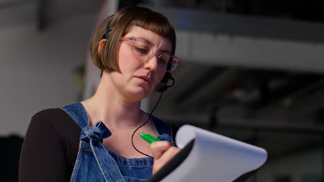 Female-Assistant-Film-Director-Wearing-Headset-With-Clipboard-Checking-Script-Shooting-Movie-Or-Video-Scene-In-Studio